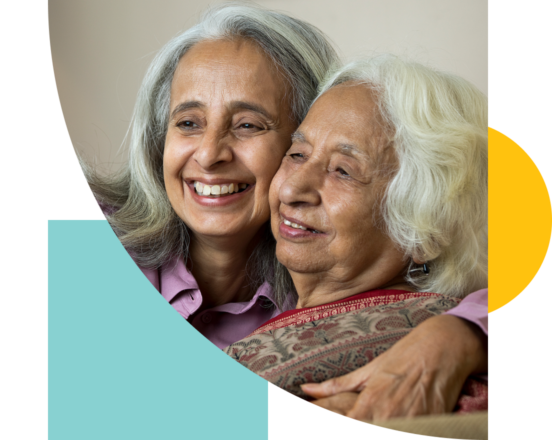 A mother and daughter of East Asian descent smiling and hugging one another. The daughter is a middle aged woman with shoulder length grey hair, while her mother is dressed in a sari and has white, wavy hair.