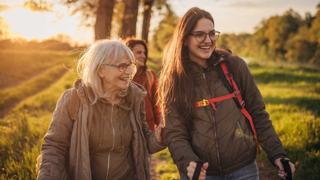 A family on a morning hike together. An older white woman with glasses walks with a smiling young woman with long brown hair. Right behind them is another woman who is out of focus.  