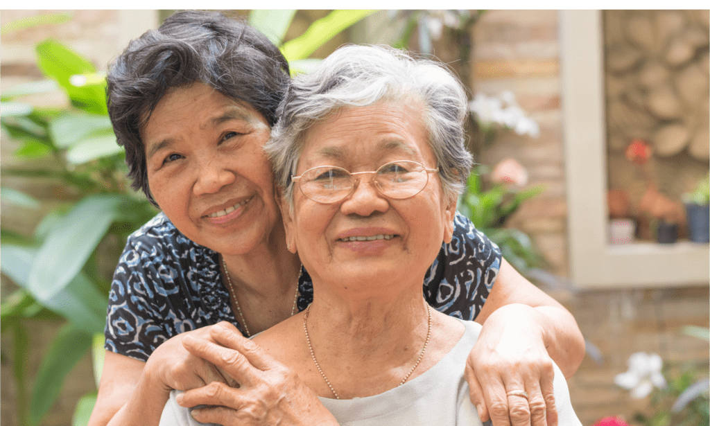 A middle-aged Asian woman with short black hair leaning over the shoulders of an elderly Asian woman with wavy silver hair who is wearing glasses.