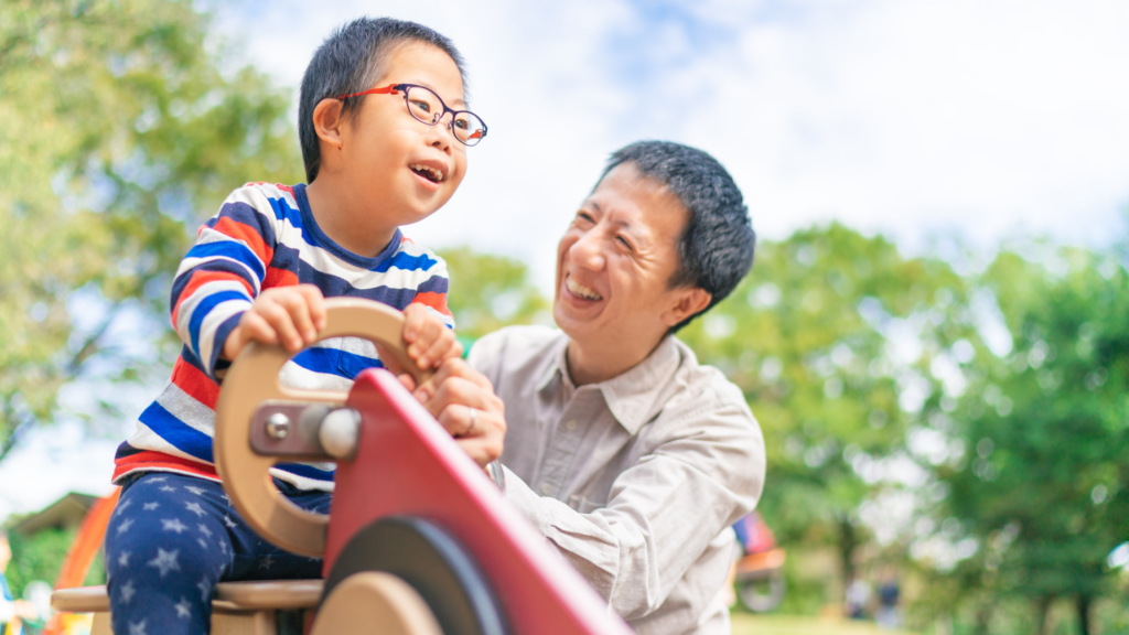 An Asian child with Down syndrome holding the wheel of a toy car. His father watches him, smiling.