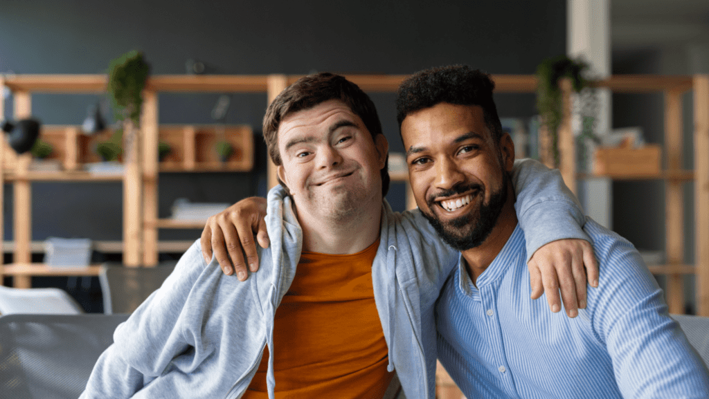 Un jeune homme blanc atteint du trisomie 21 et un jeune homme noir barbu se sourient en s'entourant de leurs bras.  