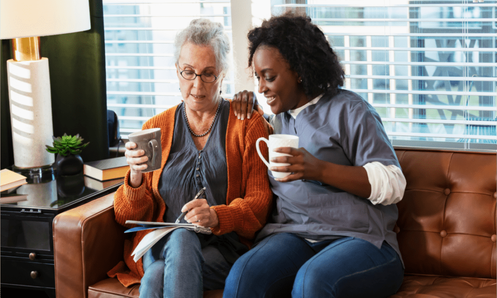 Une femme blanche âgée portant des lunettes est assise à côté d'une jeune femme noire sur un canapé. Les deux femmes tiennent des tasses de café et la personne âgée écrit sur un bloc-notes sous le regard de la jeune femme.