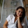 A smiling young woman with long brown hair sitting in a rattan swing chair. One hand is propping up her chin.