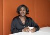 Portrait of Juanita Forde. A Black woman with a bob seated at a table holding a cup, against an orange backdrop.