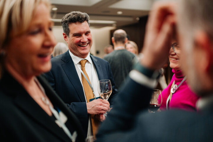 Un homme souriant tenant un verre de vin discute avec d'autres participants.