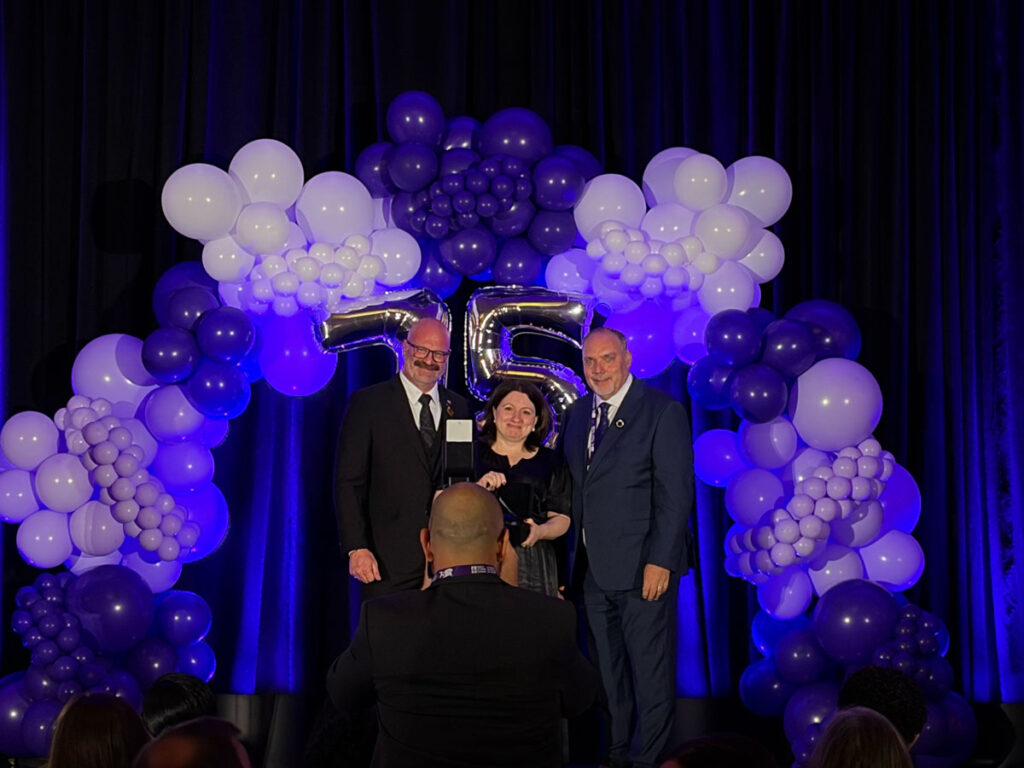 CCCE's Liv Mendelsohn on stage accepting the Changemaker award, along with the presenters. Behind them are clusters of purple balloons.